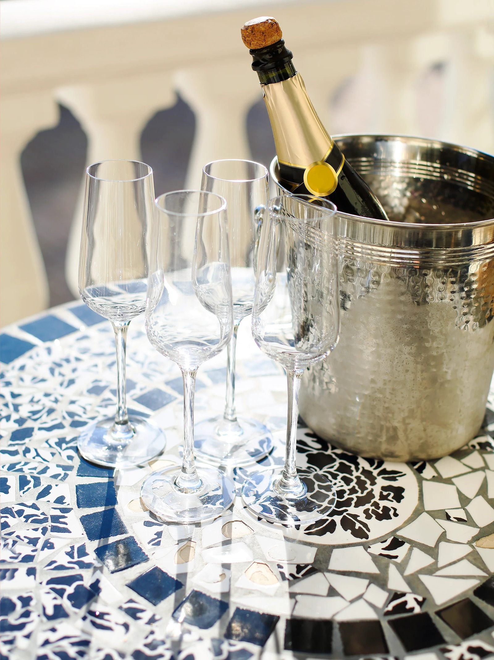 Bottle of champagne in a silver cooler with wine glasses on a mosaic table.